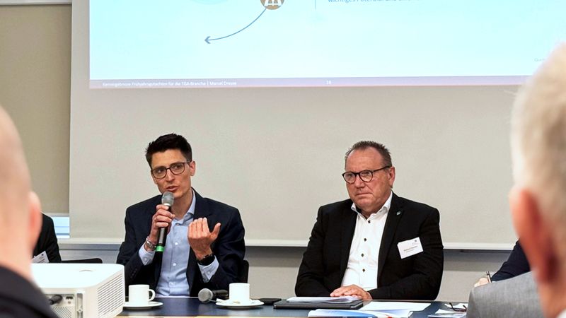 Ein Diskussionspanel bei einer Konferenz, bei dem ein Mann mit Mikrofon spricht und ein anderer aufmerksam zuhört.