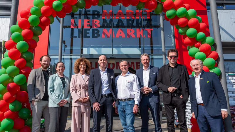 Eine Gruppe von Menschen steht vor einem Eingang, der mit einem Bogen aus roten und grünen Luftballons dekoriert ist.