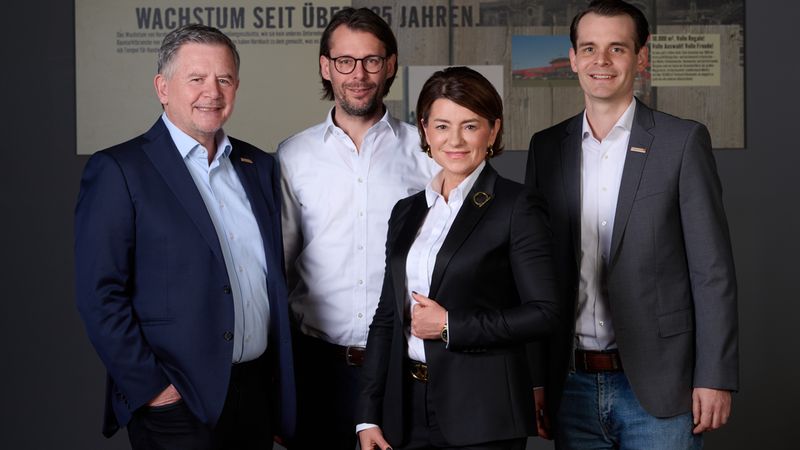 Ein Gruppenfoto von vier Geschäftsleuten in formeller Kleidung vor einem Hintergrund mit Text.