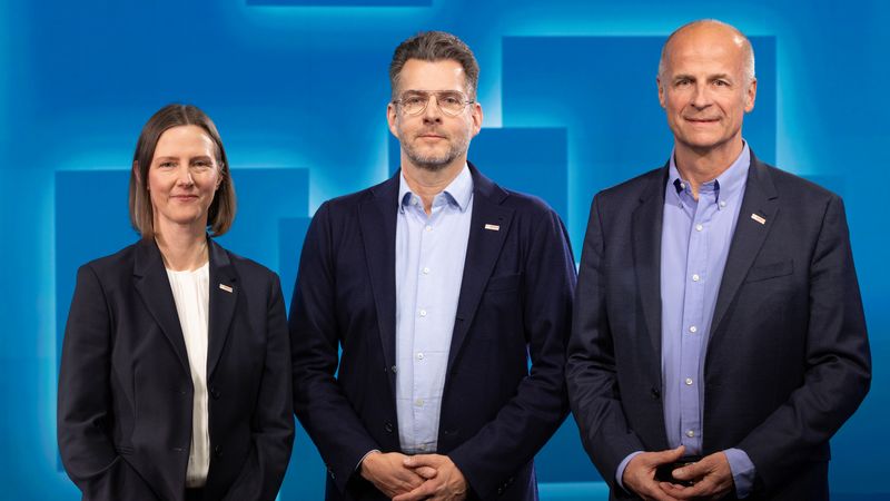 Ein Gruppenfoto von drei Personen in formeller Kleidung vor einem blauen Hintergrund.