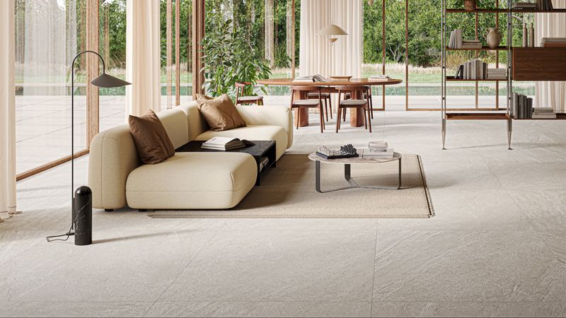 Ein modernes Wohnzimmer mit einem großen Fenster, das viel natürliches Licht hereinlässt. Es gibt ein beiges Sofa, einen Couchtisch und einen Esstisch im Hintergrund.