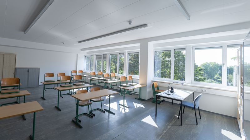 Ein leerer Klassenraum mit mehreren Tischen und Stühlen, großen Fenstern und viel natürlichem Licht.