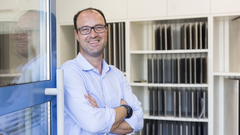 Ein Mann steht lächelnd in einem modernen Büro mit Regalen im Hintergrund.