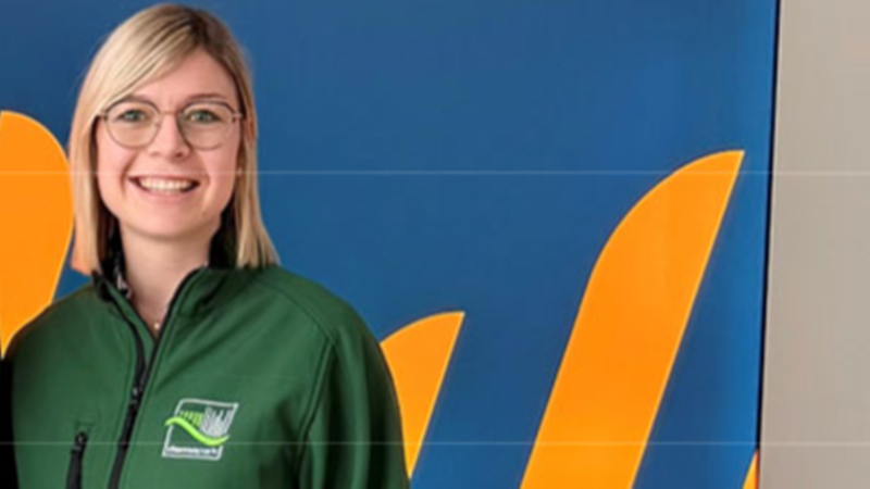 Eine Frau mit Brille und grüner Jacke steht vor einem bunten Hintergrund.