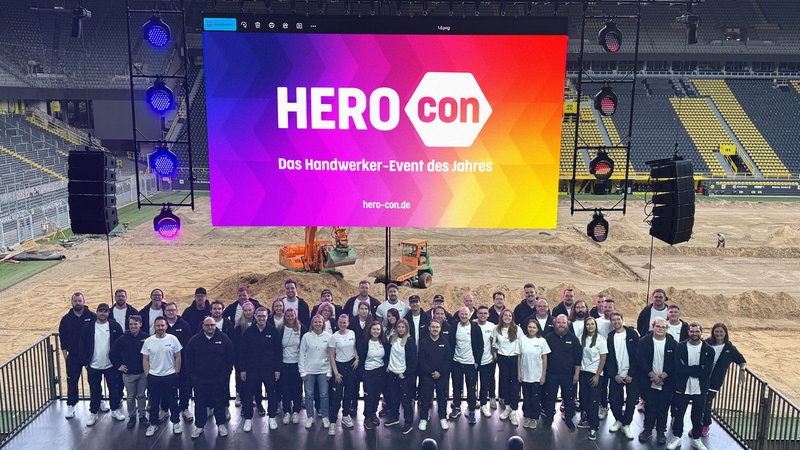 Gruppe von Menschen vor einer großen Bühne im Stadion mit einem leuchtenden Bildschirm, der das Handwerker-Event des Jahres ankündigt.