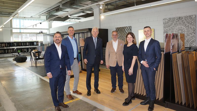 Eine Gruppe von sechs Personen in formeller Kleidung steht in einem modernen Ausstellungsraum mit Bodenbelägen und Mustern.