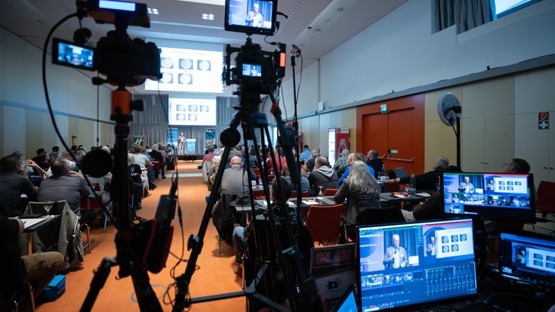 Ein Konferenzraum mit mehreren Kameras und Teilnehmern, die einer Präsentation folgen.