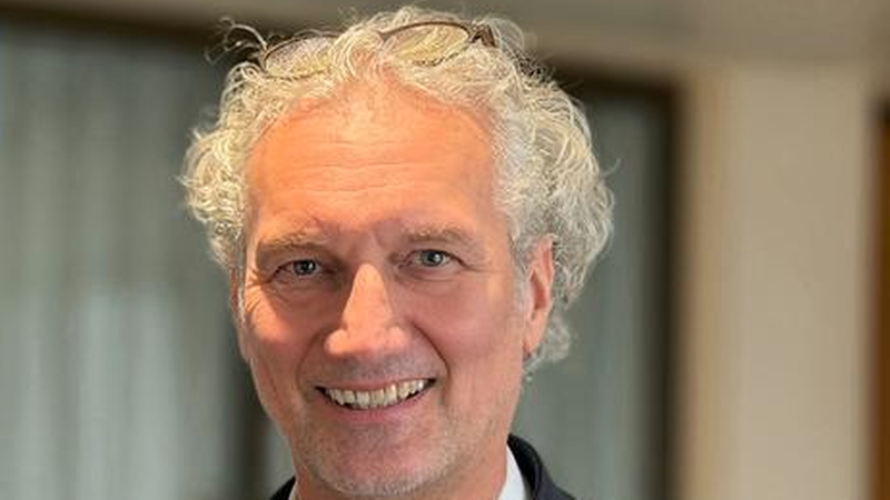 Ein eleganter Mann mit grauem Haar und einem freundlichen Lächeln in einem dunklen Anzug.