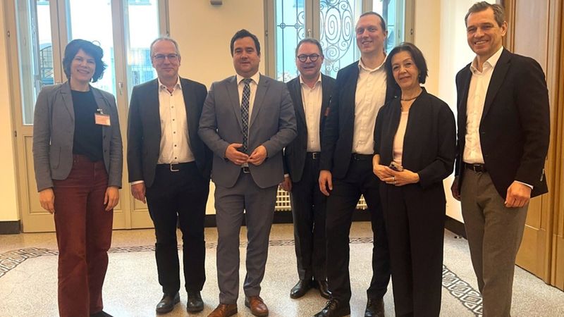 Eine Gruppe von sieben Personen steht in einem Raum vor großen Fenstern und lächelt in die Kamera.