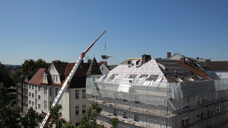 Ein Kran hebt Baumaterialien auf das Dach eines mehrstöckigen Gebäudes, während Arbeiter an der Dachrenovierung arbeiten.