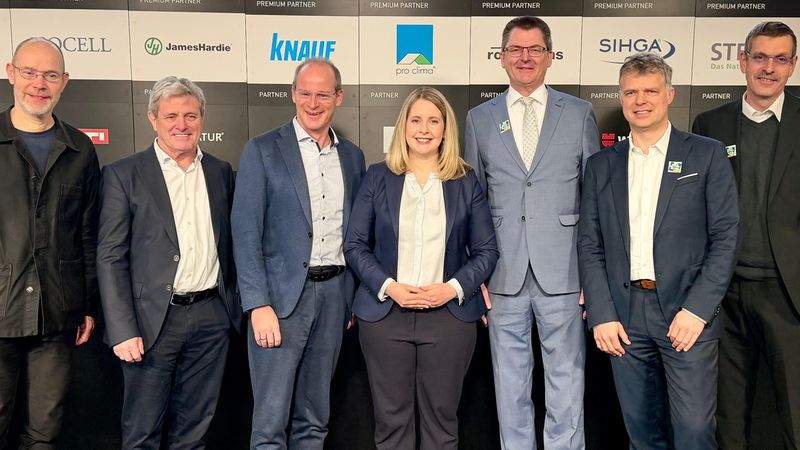 Eine Gruppe von sieben Personen steht vor einer Sponsorenwand und posiert für ein Foto.