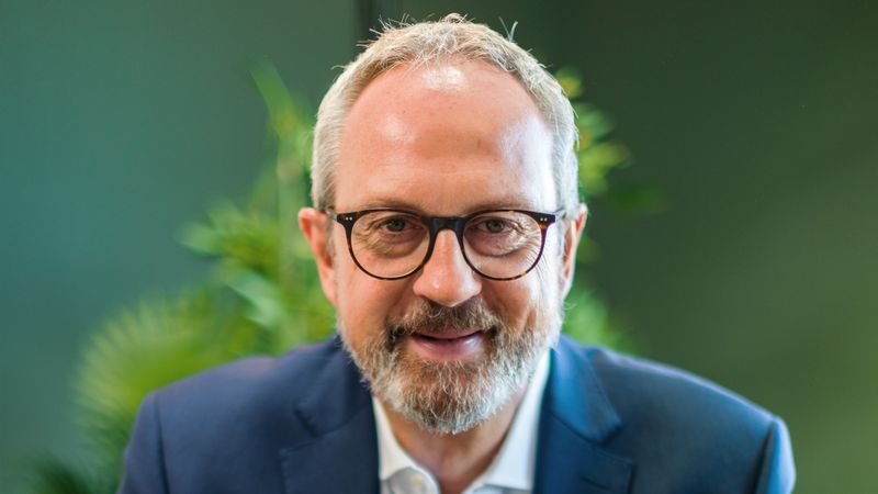 Ein Mann in einem blauen Anzug sitzt lächelnd vor einer grünen Pflanze.