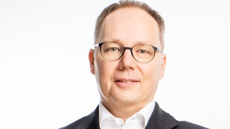 Ein Mann mit Brille in einem schwarzen Anzug vor einem weißen Hintergrund.