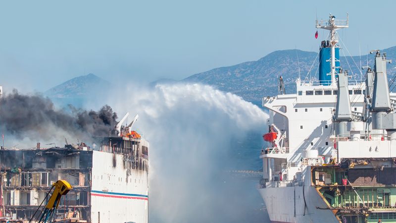 Ein Schiff löscht einen Brand auf einem anderen Schiff im Hafen mit einem starken Wasserstrahl.