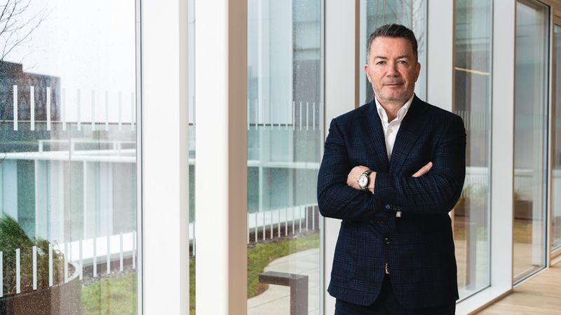 Ein Mann in einem dunklen Anzug steht mit verschränkten Armen vor einem großen Fenster in einem modernen Gebäude.