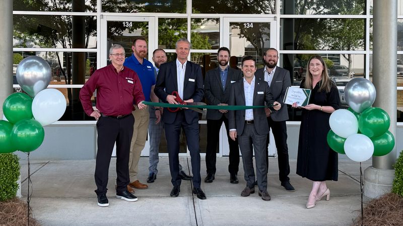 Eine Gruppe von Menschen steht vor einem Gebäude und schneidet ein grünes Band durch, um eine Eröffnung zu feiern. Luftballons in Grün und Silber schmücken den Eingang.