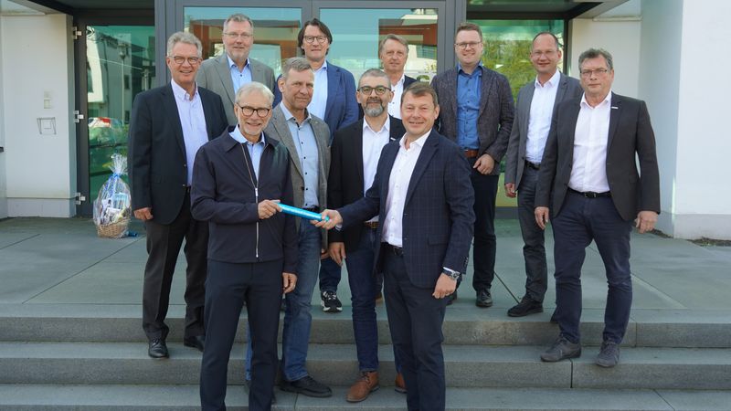 Eine Gruppe von Männern steht auf einer Treppe vor einem Gebäude, wobei zwei von ihnen einen Stab übergeben.