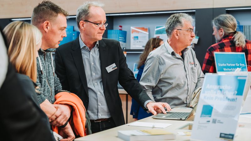 Menschen im Gespräch an einem Messestand mit Informationsmaterialien.