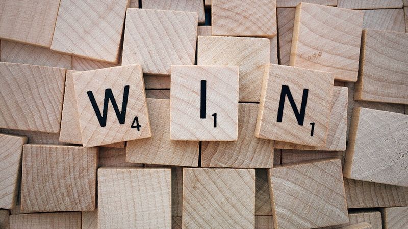 Ein Bild von Scrabble-Buchstaben, die das Wort "WIN" bilden, auf einem Haufen von leeren Holzplättchen.