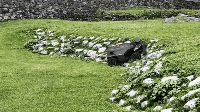 Ein autonomer Mähroboter fährt über einen gepflegten, grünen Rasen an einer steinernen Böschung; im Hintergrund ist eine niedrige Trockenmauer und üppige Vegetation zu sehen.