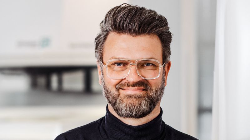 Ein Mann mit Brille und Bart steht mit verschränkten Armen in einem modernen Innenraum.