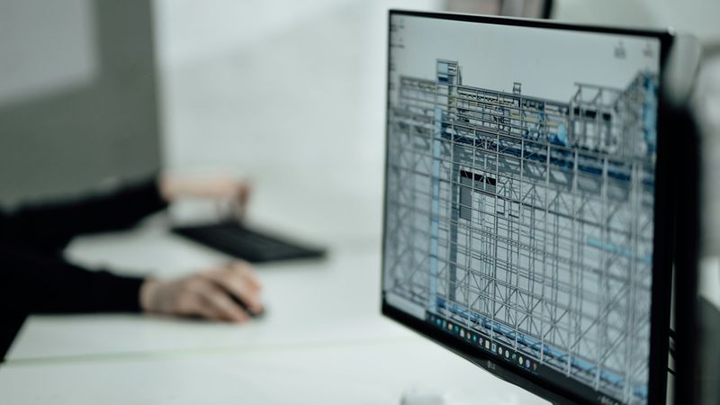 Ein Computerbildschirm zeigt ein Architekturdesign, während eine Person im Hintergrund an einer Tastatur arbeitet.