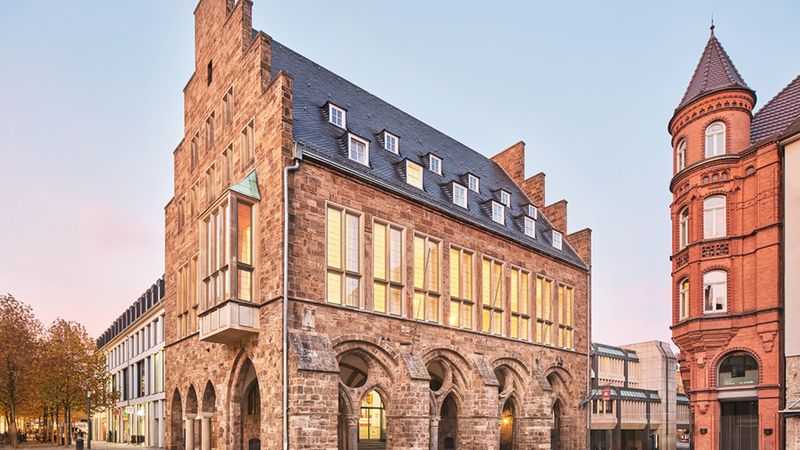 Ein historisches Gebäude aus Stein mit großen Fenstern und einem markanten Dach im Abendlicht.