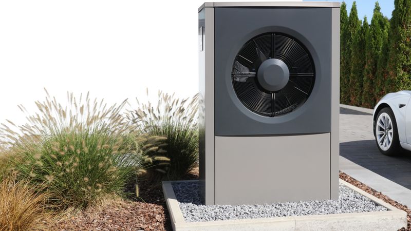 Eine Wärmepumpe im Garten neben einem modernen Auto, umgeben von Pflanzen und Kies.