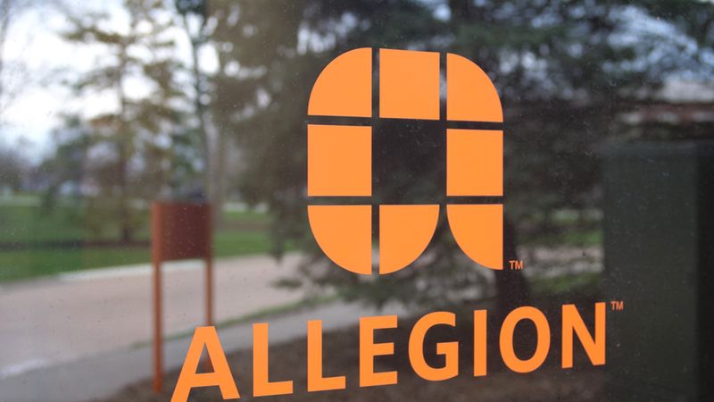 Das Bild zeigt das Allegion-Logo auf einem Fenster, mit einem verschwommenen Hintergrund von Bäumen und einem Gehweg.