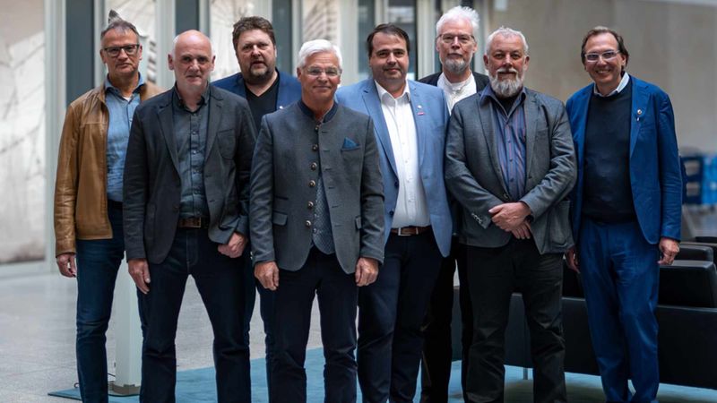 Eine Gruppe von acht Männern in formeller Kleidung steht in einem modernen Gebäude.