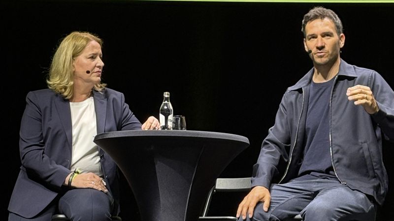 Zwei Personen sitzen auf einer Bühne an einem Tisch und diskutieren miteinander.