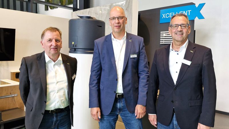 Drei Männer in Anzügen stehen vor einer modernen Wärmepumpe in einem Ausstellungsraum.