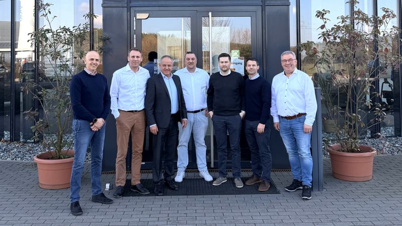 Eine Gruppe von sieben Personen steht vor dem Eingang eines Gebäudes mit dem Schriftzug "KRAMP".