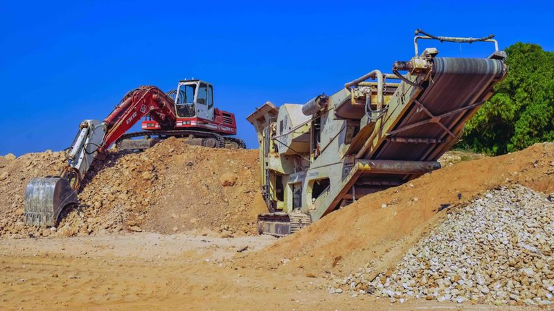 Ein Bagger und ein Förderband auf einer Baustelle, umgeben von Erdhaufen und Schutt.