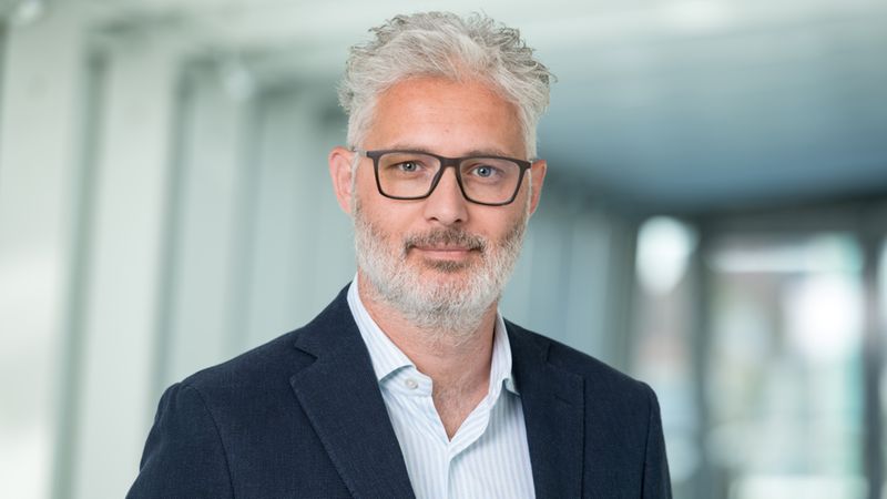 Ein Porträt eines Mannes mit grauen Haaren und Brille in einem Anzug, der in die Kamera schaut.