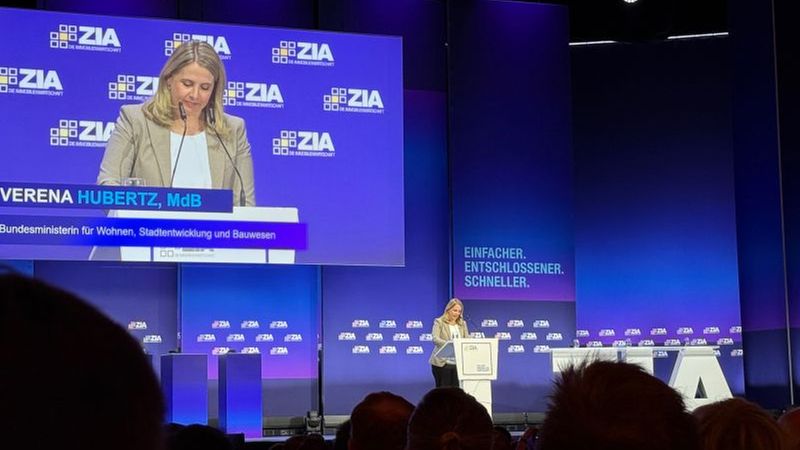 Eine Frau hält eine Rede auf einer Konferenzbühne mit einem großen Bildschirm im Hintergrund.