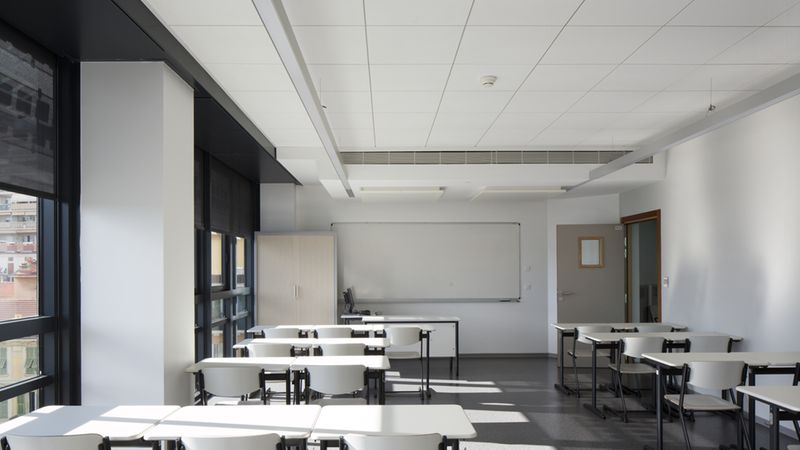 Ein leeres Klassenzimmer mit weißen Tischen und Stühlen, großen Fenstern und einer Tafel an der Wand.