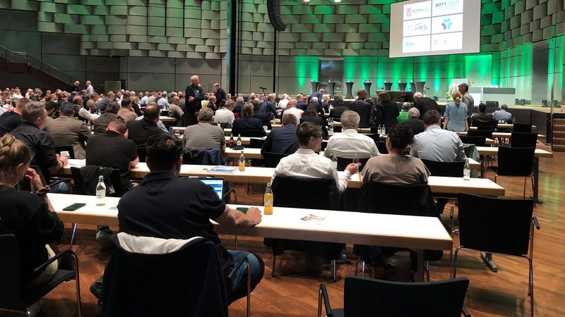 Ein großer Konferenzsaal mit vielen Menschen, die an Tischen sitzen und einer Präsentation folgen.