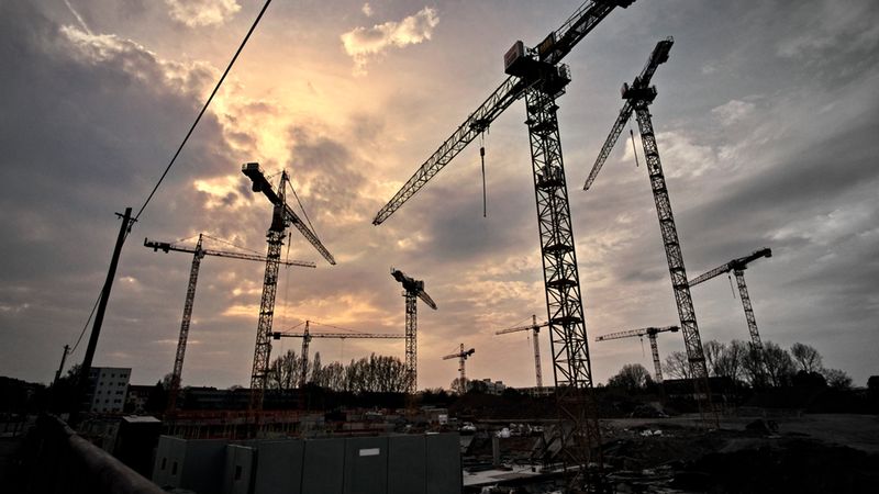 Eine beeindruckende Landschaft mit mehreren Baukränen vor einem dramatischen Sonnenuntergang.