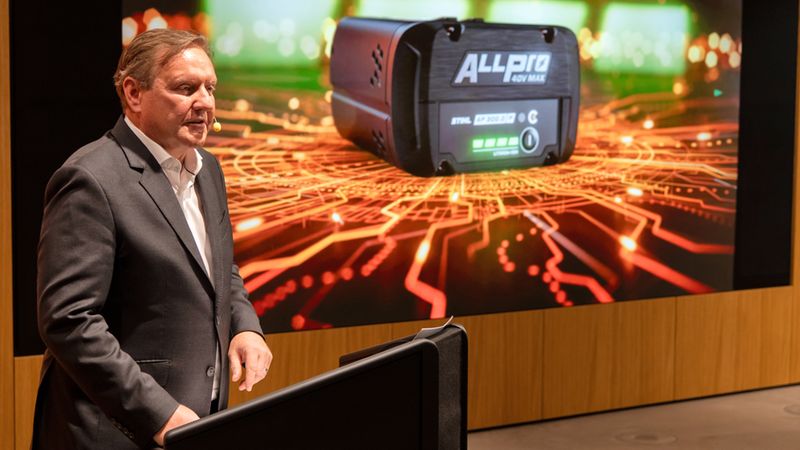 Ein Mann hält eine Präsentation über eine neue Akku-Innovation, die auf einem Bildschirm im Hintergrund zu sehen ist.