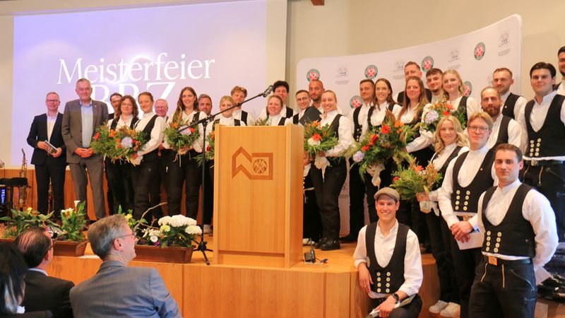 Eine Gruppe von Handwerkern in traditioneller Kleidung steht auf einer Bühne während einer Meisterfeier. Sie halten Blumensträuße und lächeln in die Kamera.