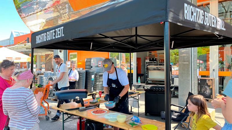 Eine Grillvorführung im Freien vor einem Baumarkt, bei der ein Grillmeister Speisen zubereitet und von einer kleinen Gruppe von Menschen beobachtet wird.