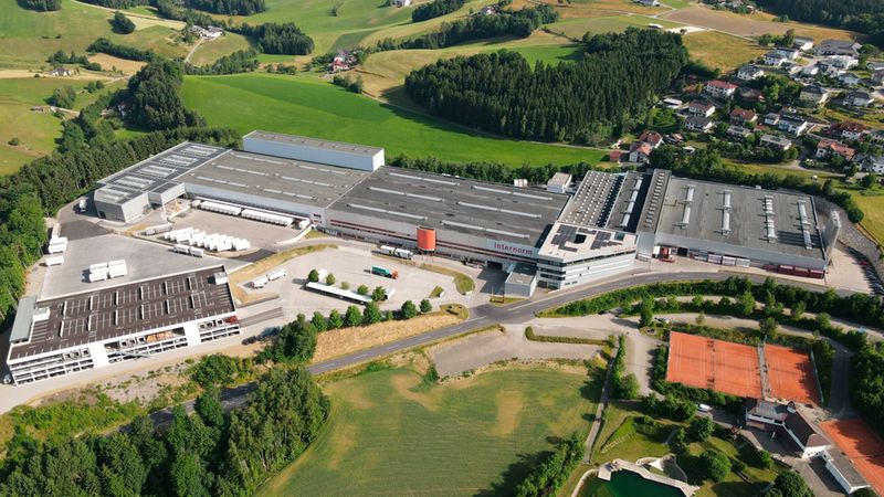 Luftaufnahme eines großen Fabrikkomplexes inmitten einer grünen Landschaft mit umliegenden Feldern und Wäldern.