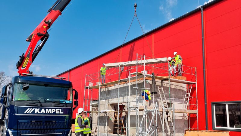 Ein Kran hebt ein Bauteil auf einer Baustelle an, während Bauarbeiter auf einem Gerüst arbeiten. Im Hintergrund ist ein rotes Gebäude zu sehen.