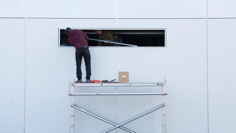 Ein Arbeiter steht auf einem Gerüst und arbeitet an einem Fenster in einer weißen Wand.