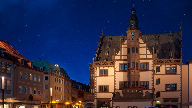 Das Bild zeigt ein historisches Rathaus bei Nacht, umgeben von beleuchteten Gebäuden und einem klaren Sternenhimmel.