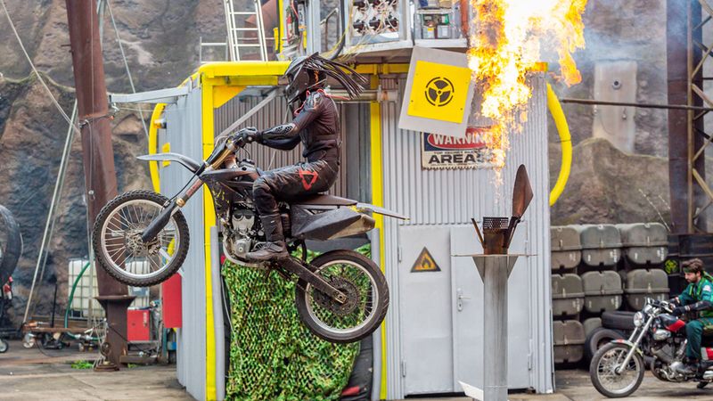 Ein Motorradfahrer führt einen spektakulären Stunt durch, indem er über eine Rampe springt, während Flammen in die Luft schießen.