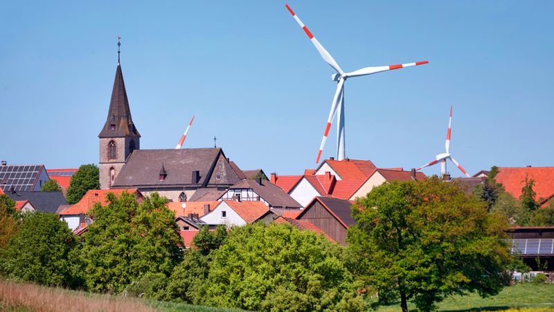 Ein idyllisches Dorf mit einer Kirche im Vordergrund und mehreren Windkraftanlagen im Hintergrund unter klarem Himmel.