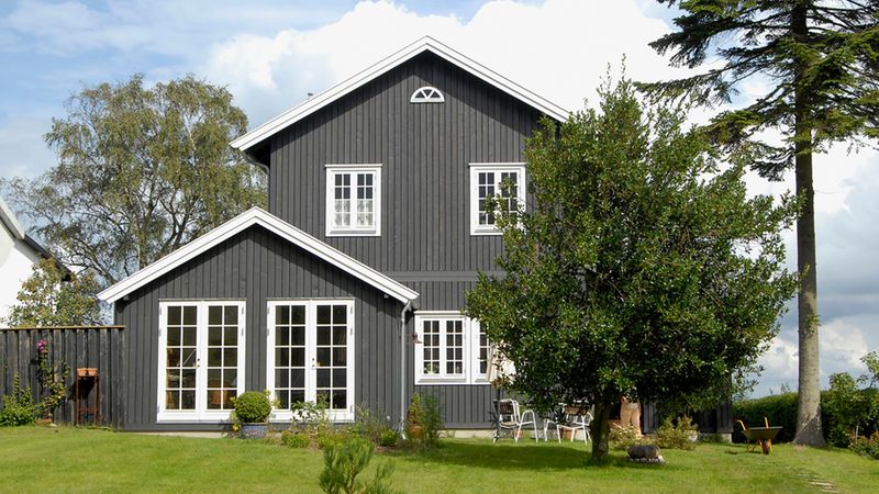 Ein modernes graues Holzhaus mit großen Fenstern, umgeben von einem gepflegten Garten mit Bäumen und Rasen.