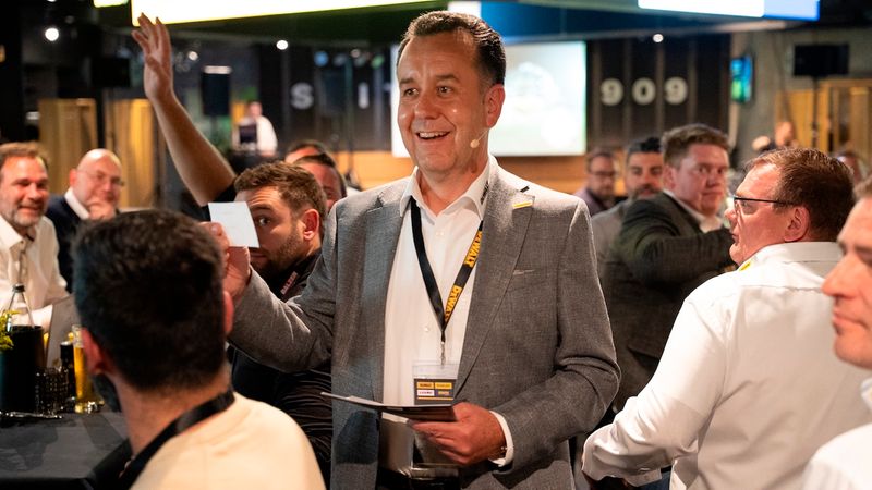 Ein Mann in einem grauen Anzug hält eine Karte in der Hand und lächelt, während er bei einem Event mit mehreren Personen im Hintergrund steht.
